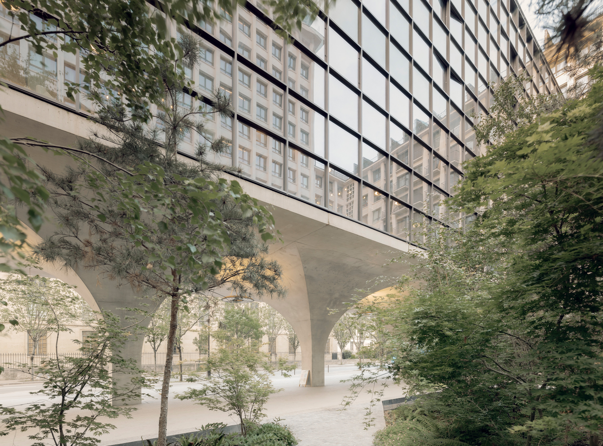 Landscaped courtyard at Morland Mixité Capitale, where planted gardens and mature trees extend beneath an elevated glazed volume supported by concrete arches, integrating nature and public space within the transformed urban block.