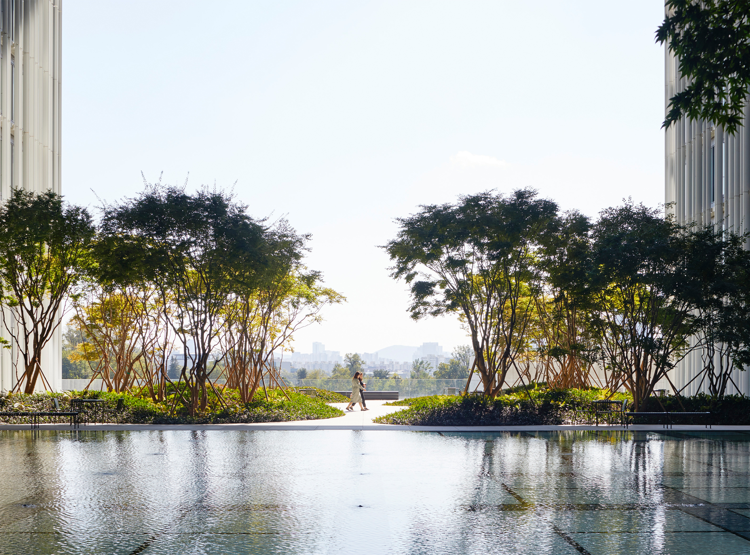 Landscaped courtyard at the Amorepacific headquarters featuring a shallow water terrace reflecting the sky, framed by trees and views towards the city beyond.